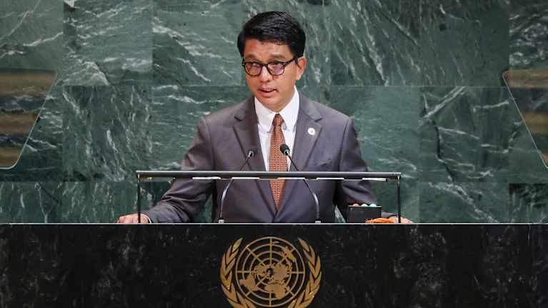 Mies puvussa puhuu pöydän takana. Pöydässä on YK:n logo. 