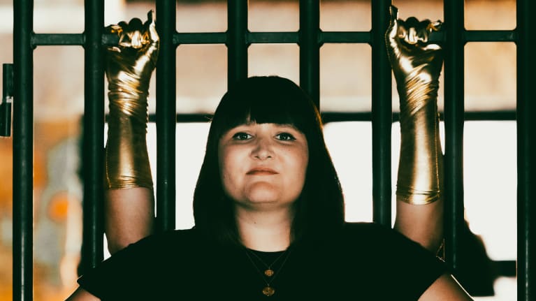 Anna Brotkin seisoo kädet länteilla kalterioven edessä ja hänen takanaan kaksi kultahanskaista kättä pitää kaltereista kiinni.