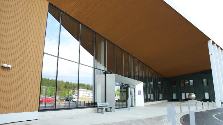 Sairaalarakennuksen päivystyksen potilasisäänkäynti jossa puurakennetta, valkoisia tolppia ja iso lasinen seinä, etualalla jalkakäytävää, matalia tolppia erottamasta autotien ja penkki.
