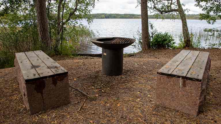 ulkoilureitin metallinen nuotiopaikka kivisine penkkeineen. Taustalla näkyy lampi