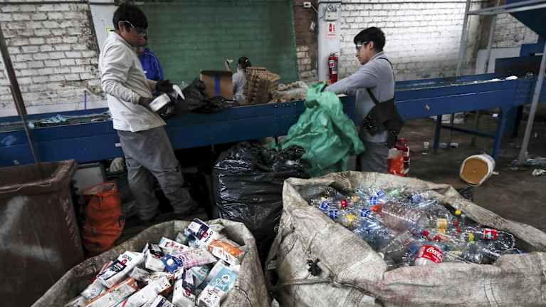 Jätteenlajittelijoita ja jätesäkkejä. Toisessa säkissä on kartonkipakkauksia, toisessa muovipulloja.