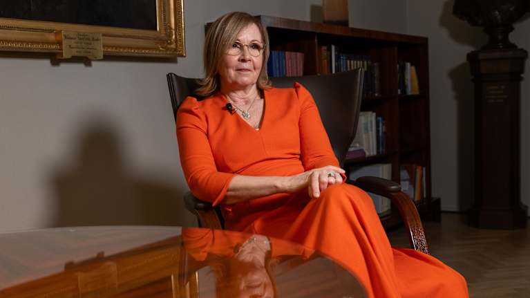 Nainen istuu oranssissa mekossa nojatuolissa sisätiloissa; taustalla kirjahylly ja taulu.