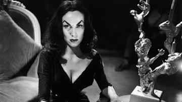 Nainen goottilaisessa mekossa istuu kynttilänjalalla koristellun arkun ääressä, pöydässä kaiverrettuna englanniksi sanat "REST IN PEACE".