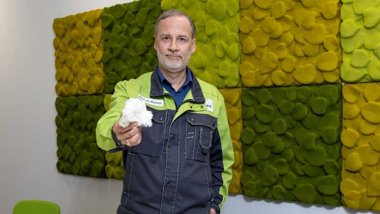 Mies esittelee käsissään olevan uudenlaisen kuitutuotteen modernissa toimistoympäristössä, taustallaan vihreän sävyinen akustiikkapaneeli.