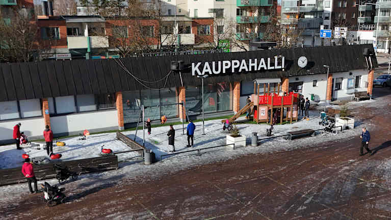 Ilmakuvassa on talvinen näkymä torialueella sijaitsevasta Lappeenrannan Kauppahallista, jonka edustalla on puistoalue, jossa lapset leikkivät lumessa.