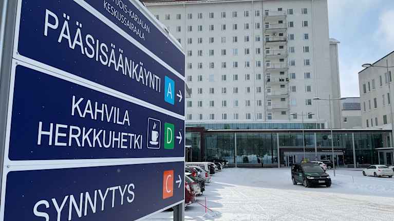 Pohjois-Karjalan keskussairaala talvisessa maisemassa: Lunta sataa kevyesti sisäänkäynnin edustalla, jossa opasteet ohjaavat pääsisäänkäynnille, kahvilaan ja synnytysosastolle.