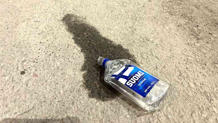 Tyhjä Suomiviina-pullo lojuu asvaltin pinnalla, jättäen varjon alustaansa.