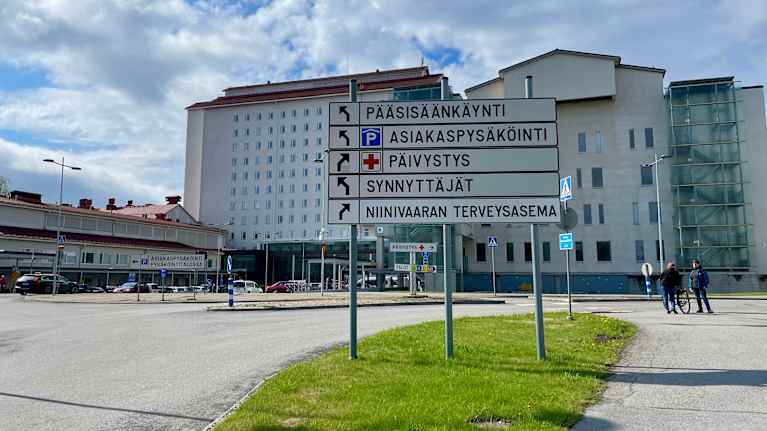 Kuvassa on sairaalan päärakennus, jonka edessä on opastekyltti. Opastekyltti ohjaa pääsisäänkäynnille, asiakasparkkiin, päivystykseen, synnytysten osastolle ja Niinivaaran terveysasemalle. Aurinko paistaa osittain pilvien takaa, ja ihmisiä näkyy kulkemassa sairaalan edustalla.