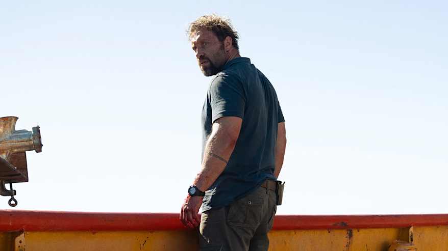 En man står på ett båtdäck och ser förvånad ut, relingen och däcket är väldigt blodigt och vid sidan syns två människoben som är delvis upprivna.