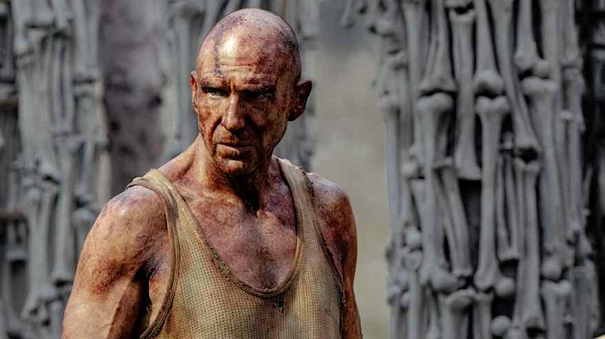 En bister Ralph Fiennes insmord i rödaktig olja står framför en mur av ben.