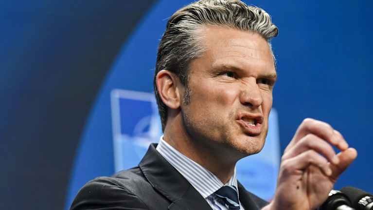 Peter Hegseth puhyy mikrofoniin oikeaa kättään nostaen. Hänellä on tumma puku, raidallinen kauluspaita ja kravatti. Tausta on sininen.