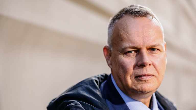 Mies seisoo pukeutuneena siniseen pukuun ja solmioon, yllään tumma päällystakki, ulkona rakennuksen edustalla.