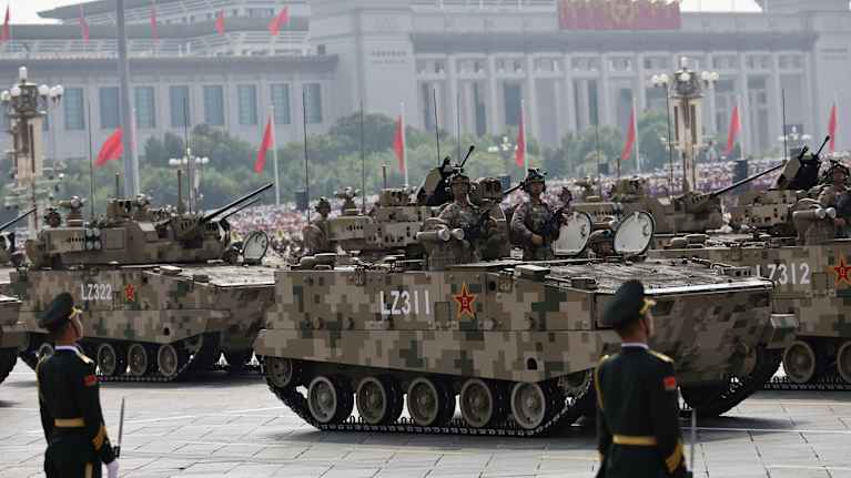 Kiinan armeijan panssaroidut ajoneuvot osana sotilasparaatia Pekingissä Taivaallisen rauhan aukiolla. Taustalla näkyy kansallisia rakennuksia ja punaisia lippuja liehuu kaikkialla.