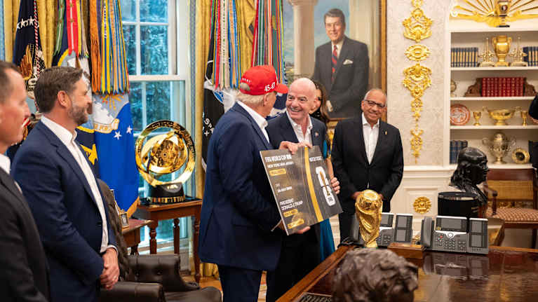 Yhdysvaltain presidentti Donald Trump pitelee työhuoneessaan kädessään suurta pahvista pääsylippua ja katsoo Fifan puheenjohtaja Gianni Infantinoa. Pöydällä on Fifan kultainen pokaali ja taustalla toinen Fifan kultainen pokaali.