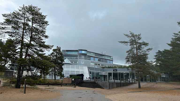 Harmaa ja lasinen rakennus kohoaa taustalla, etualla hienon hiekan alle osittain peittynyt asfaltoitu kulkuväylä.