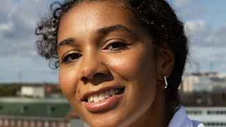 Henkilö poseeraa ulkona kirkkaassa päivävalossa valkoinen paita yllään. Taustalla näkyy kaupunkimaisema ja sininen taivas pilvillä.