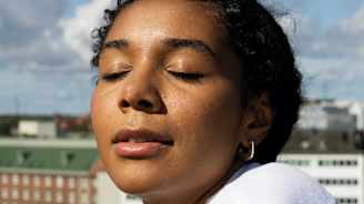 Henkilö pukeutuneena valkoiseen paitaan seisoo korkealla paikalla silmät kiinni, taustallaan kaupungin rakennukset ja taivaalla pilviä.