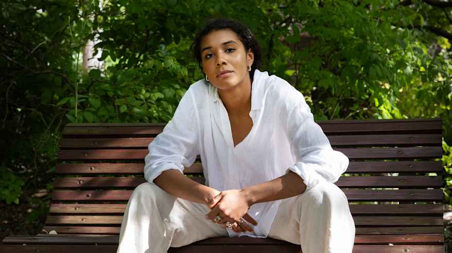 Henkilö istuu penkillä puistossa pukeutuneena valkoiseen puseroon ja vaaleisiin housuihin. Aurinko paistaa ympäröivään vehreyteen.