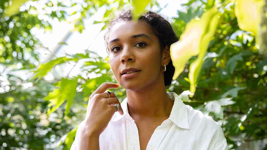 Vihreän kasvillisuuden ympäröimä henkilö pukeutuneena valkoiseen kauluspaitaan seisoo ulkona, nojaten yhdellä kädellä leukaan.