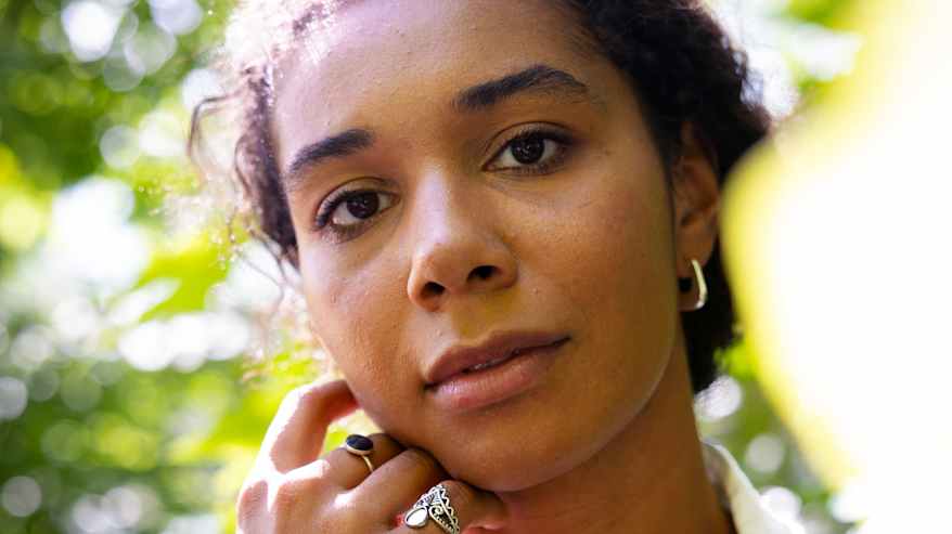 Henkilö poseeraa vehreässä ulkoilmamiljöössä pukeutuneena rentoon, valkoiseen paitaan.