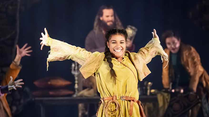 Keskellä näyttämöä näyttelijä ilmentää voimakasta liikettä taustallaan hämäriä lavasteita ja muita esiintyjiä.