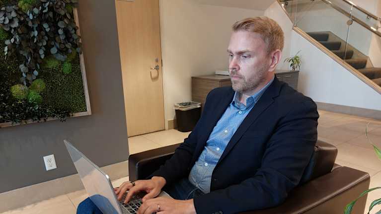 Mies istuu toimistossa kannettava tietokone sylissään.