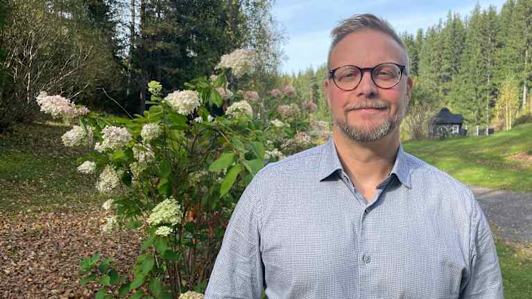 Mies seisoo syksyisessä maisemassa, taustalla vihreitä puita ja kukkiva syyshortensia.