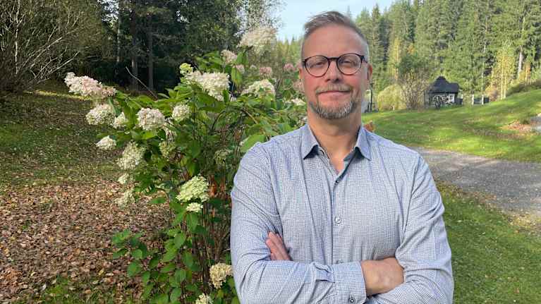 Silmälasipäinen mies seisoo kädet eteensä ristittyinä kauniissa syksyisessä maisemassa, taustalla vihreitä puita ja kukkiva syyshortensia.