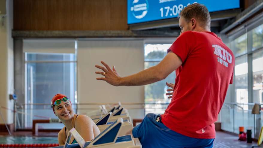 Valmentaja ohjeistaa uimaria altaalla innokkaasti. Taustalla näkyy kellotaulu, joka näyttää ajan ja päivämäärän.