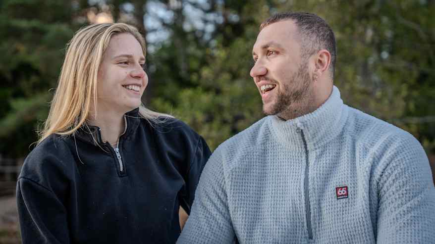 Kaksi henkilöä istuu ulkona syksyisessä maisemassa, pukeutuneina lämpimiin vaatteisiin.