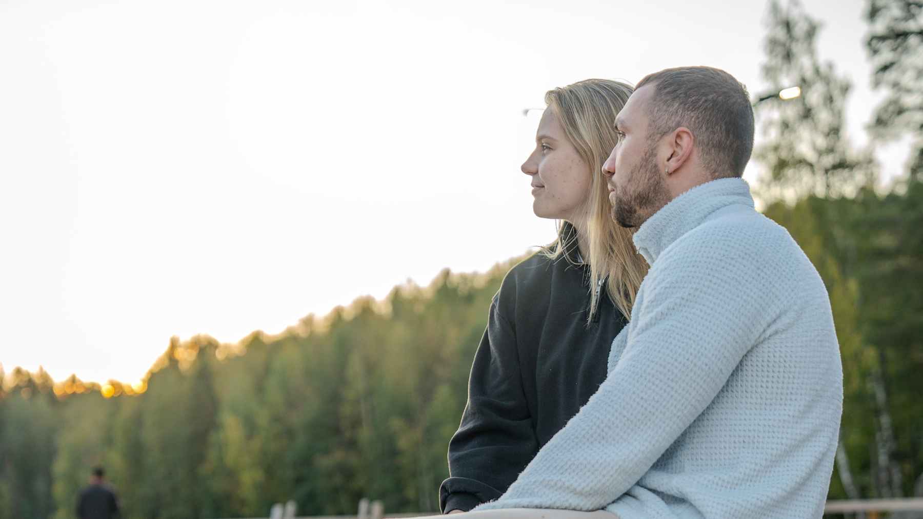 Kaksi ihmistä istuu laiturilla järven rannalla aurinkoisena iltana. Taustalla näkyy metsä ja rauhallinen järvimaisema.