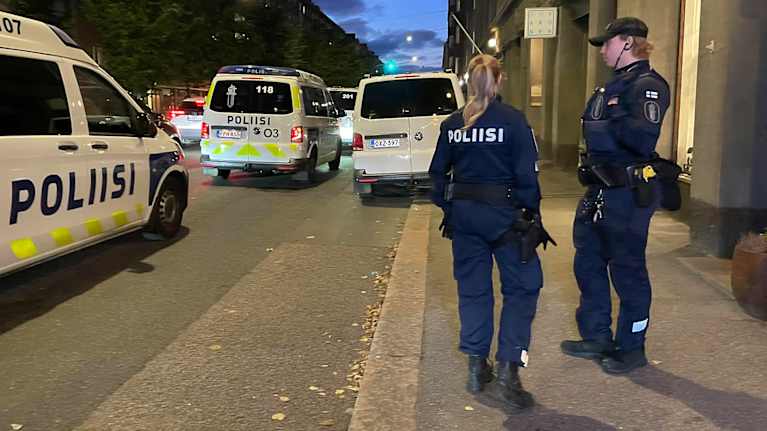 Kuvassa näkyy kaksi poliisia seisomassa kadulla, ja taustalla on useita poliisiautoja, jotka ovat parkkeerattu tien varteen.