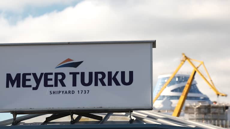 Meyer Turun telakan logo kyltissä telakan pääportilla. Taustalla näkyy telakan nosturi ja valmisteilla olevan aluksen yläosia.