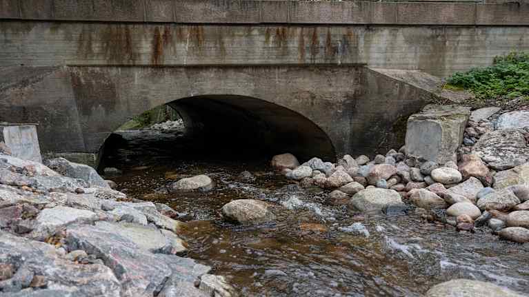 Betoninen silta, jonka alta kulkee oja. Ojan reunoilla näkyy leikkauskohdat, joista ennen lähti pato.