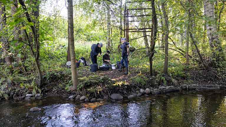 Neljä miestä vehreällä ojanpenkalla. Yksi mies polvillaan maassa ämpäreiden ja laatikon ääressä.