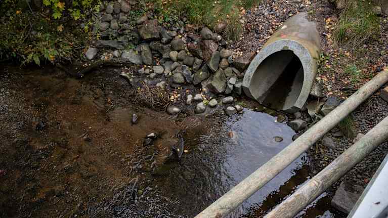 Betoninen sadevesiviemäriputken pää, josta tulee vettä ojaan. Aukon vieressä vaaleampaa ja hienompaa hiekkaa joen pohjassa.