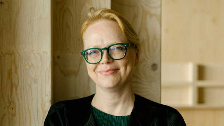 Henkilö seisoo puisella taustalla pukeutuneena mustaan takkiin ja vihreään puseroon, kädet ristittyinä rinnan yli.