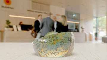 Keskellä kuvaa lasinen taide-esine. Taustalla on rakennuksen vastaanottoalue, jossa työntekijät keskustelevat toistensa kanssa.