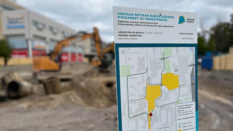 Kuvassa kyltti työmaan edessä. Kyltissä lukee mm. Vantaan ratikan rakentaminen. Taustalla auki kaivettu tie ja kaivinkone.