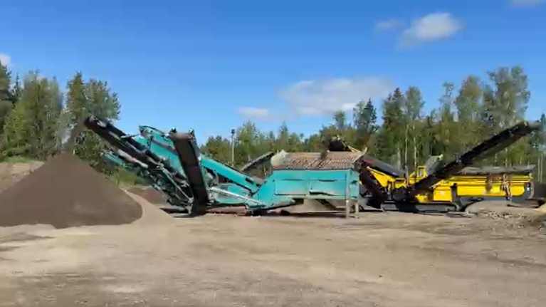 Kuvassa näkyy teollinen maankäsittelykone, joka seuloo maata ja lajittelee sen kasaan. Taustalla on metsää ja sininen taivas.