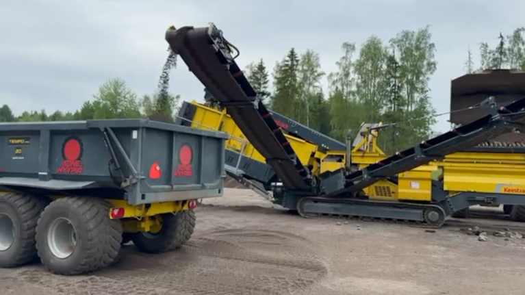 Kentuck-merkkinen seulontakone seuloo maa-ainesta ja syöttää sitä maansiirtoajoneuvon kippilavalle. Taustalla näkyy metsää ja pilvinen taivas.