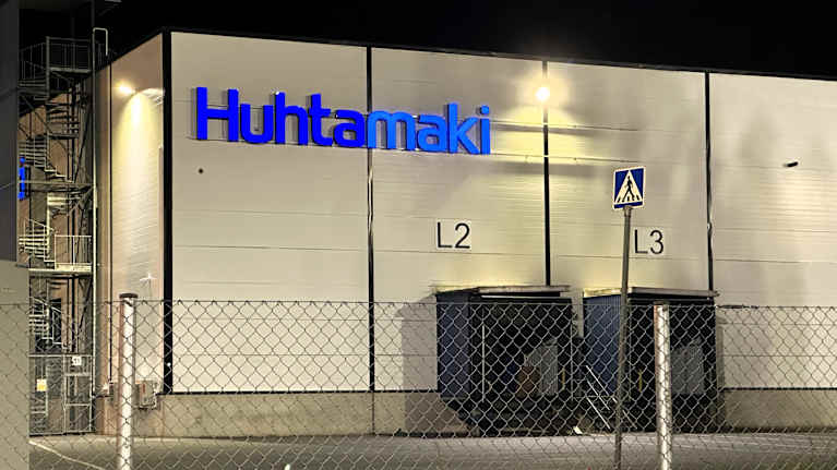 Huhtamäen tehdasrakennus yön valaistuksessa, etualalla näkyy aita ja portti.