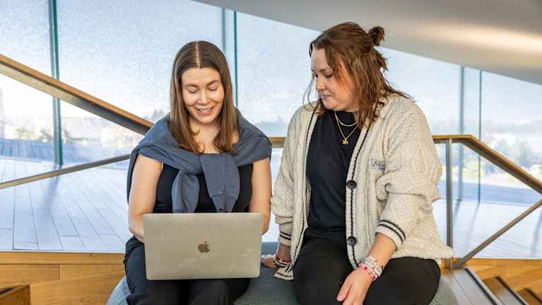 Kaksi henkilöä katselevat kannettavaa tietokonetta yhdessä modernissa ja valoisassa sisätilassa.