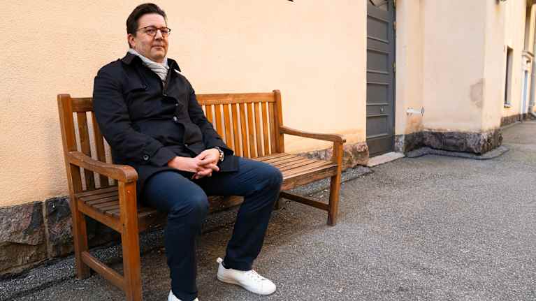 Nuori mies istuu puisella penkillä ulkona pukeutuneena paksuun takkiin ja valkoisiin kenkiin.