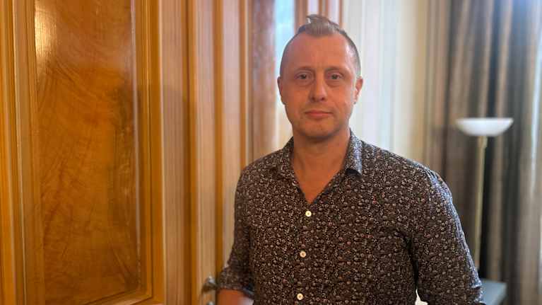 Mies seisoo huoneessa, jossa on puinen paneeliseinä ja verhot.