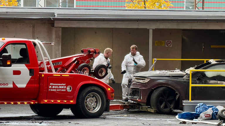 Kaksi valkoisiin haalareihin pukeutunutta miestä seisoo parkkihallin ulkopuolella. Heidän edessään on hinausauto, joka hinaa tulipalossa palanutta autoa.