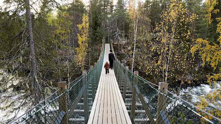 Kaksi henkilöä kävelee kosken ylittävällä riippusillalla.