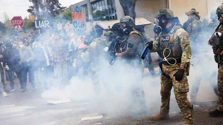 Raskaasti varustautuneita kansalliskaartin jäseniä seisoo tiellä. Taustalla näkyy mielenosoittajia, ja ilmassa on savua tai kaasua.