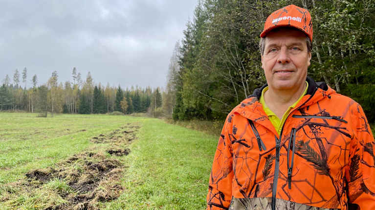 Hannu Saukkola seisoo vihreän pellon laidassa metsän reunassa. Hänen vieressään pellossa on pitkä musta kaivuujälki, joka on villisikojen tekemä. 