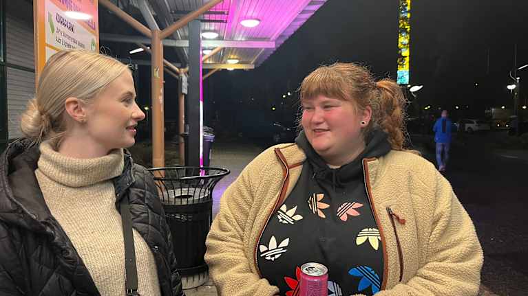 Kaksi nuorta naista ABC-huoltoaseman pihalla ilta-aikaan.